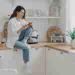 Female shopping groceries online by mobile phone app, sitting in modern kitchen at home. E commerce